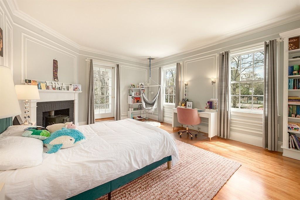 This bright and airy bedroom features light-toned walls with classic molding, hardwood flooring, and a large area rug. The room is furnished with a bed adorned with plush pillows, a desk with a pink chair, and a hanging hammock chair near the window. A fireplace with a mantel serves as a focal point, while built-in shelving and large windows provide both storage and natural light.