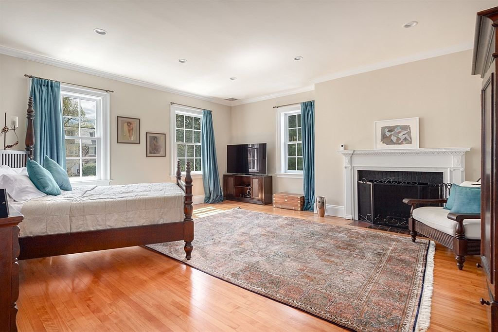 This spacious primary bedroom features warm wood flooring and a large, intricate area rug that anchors the room. A classic wooden bed frame sits to the left, while a traditional fireplace with a white mantel and a black metal screen serves as a focal point on the right. The room is brightened by three windows dressed in blue curtains, and a television is positioned on a wooden media console between two of the windows.