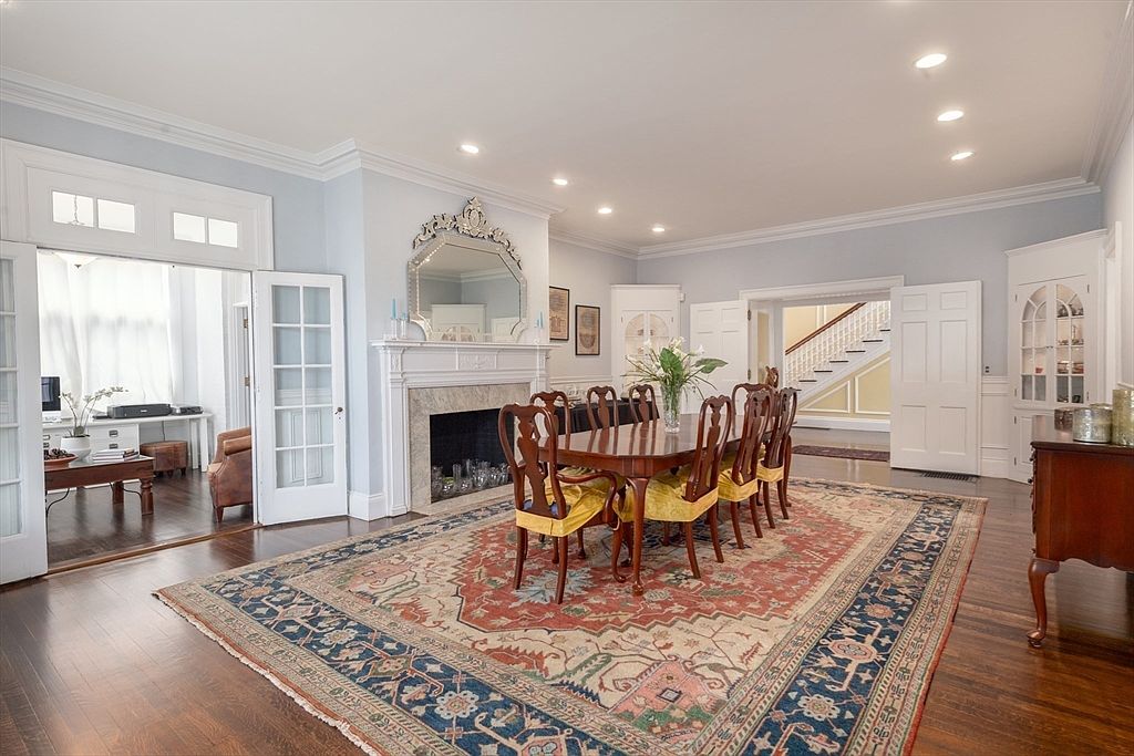 This elegant dining room features a formal wooden table surrounded by chairs with yellow upholstered seats, set atop a large, intricately patterned area rug. A prominent fireplace with a decorative mantel and a large, ornate mirror serves as the room's focal point, while built-in corner cabinetry adds a classic touch. The space is brightened by recessed lighting and flows seamlessly into an adjacent office area and a grand staircase visible through the doorway.