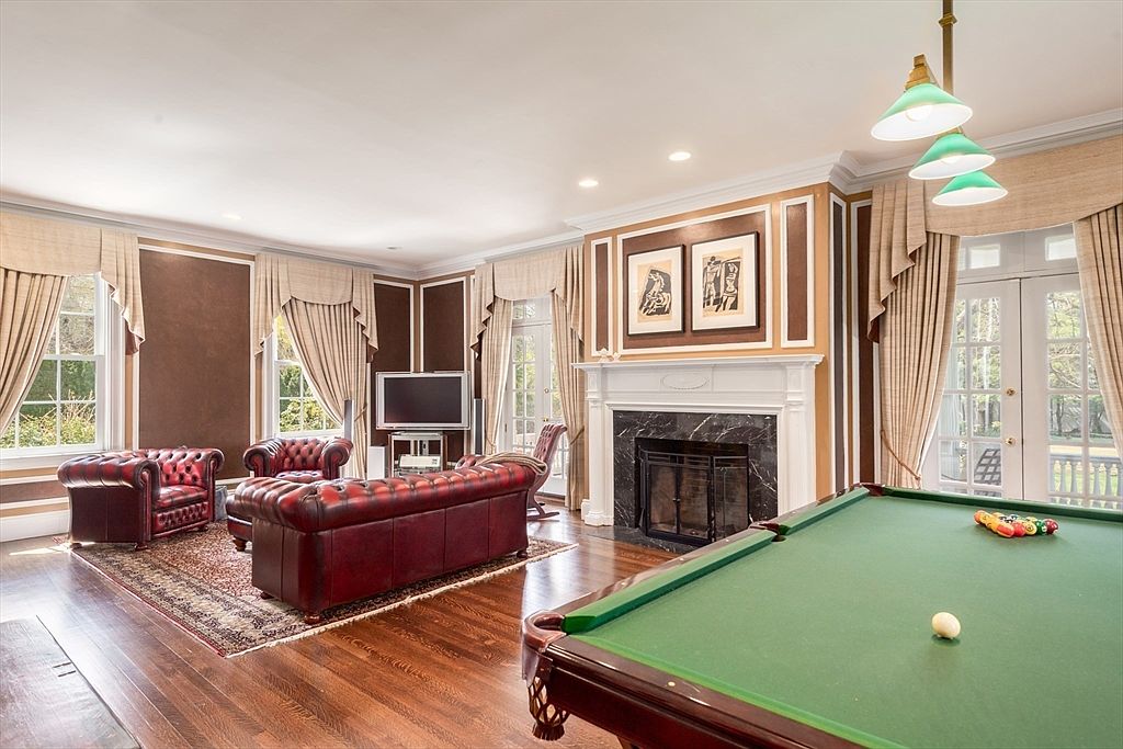 This spacious living area features a classic design with dark wood flooring, rich brown wall paneling, and a prominent fireplace with a dark stone surround and white mantel. The room is furnished with a deep red leather sofa and matching armchairs arranged on a patterned area rug, while a billiard table occupies the foreground. Large windows with elegant drapery allow natural light to fill the space, creating a sophisticated and inviting atmosphere.