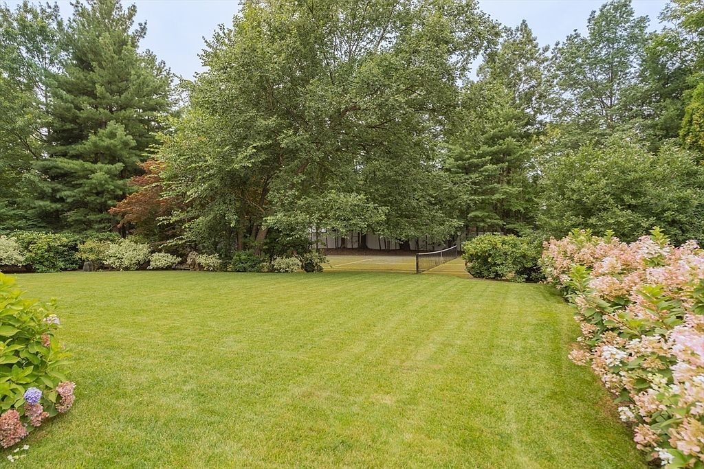 This expansive, well-manicured lawn serves as a lush backyard retreat, framed by mature trees and vibrant flowering shrubs. A tennis court with a net is visible in the background, nestled among the dense greenery. The scene offers a serene and private atmosphere, perfect for outdoor recreation and relaxation.