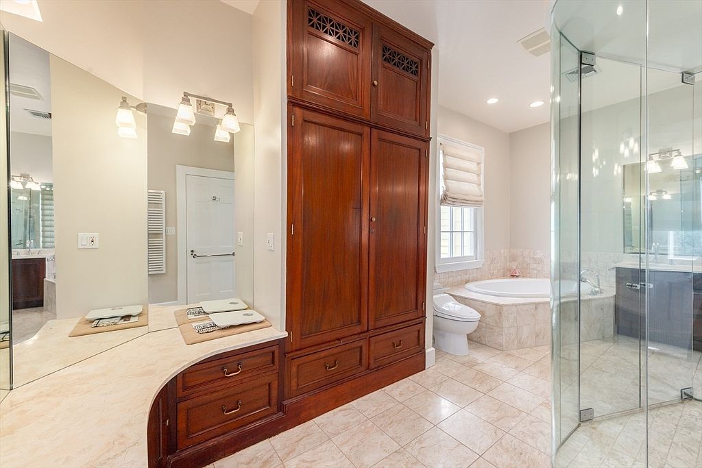 This spacious primary bathroom features a large, dark wood storage cabinet as a central focal point, flanked by a vanity area with a mirror and a glass-enclosed shower. A soaking tub is positioned in the corner near a window with a fabric shade, creating a bright and airy atmosphere. The room is finished with light-toned tile flooring and neutral wall colors, offering a clean and functional layout.