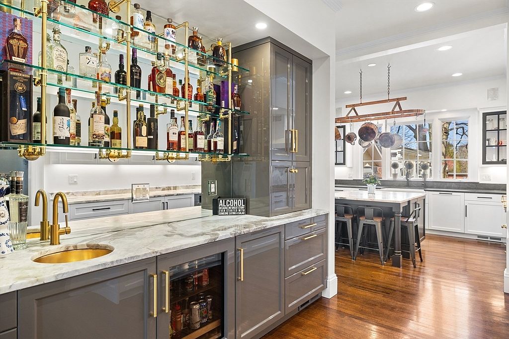 This sophisticated wet bar area features sleek grey cabinetry with gold-toned hardware and a stone countertop with an integrated sink. A multi-tiered glass shelving unit displays an extensive collection of spirits, while a small beverage refrigerator is built into the base cabinetry. The perspective looks from the bar toward an adjacent kitchen space, which includes a large island with seating and a hanging pot rack, creating a cohesive and luxurious open-concept layout.