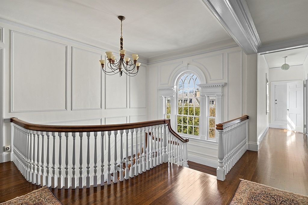 This elegant landing area features classic architectural details, including white wainscoting and a curved wooden handrail atop white balusters. A prominent arched window serves as a focal point, allowing natural light to illuminate the polished wood flooring. The space is finished with a traditional chandelier, creating a sophisticated and airy atmosphere.