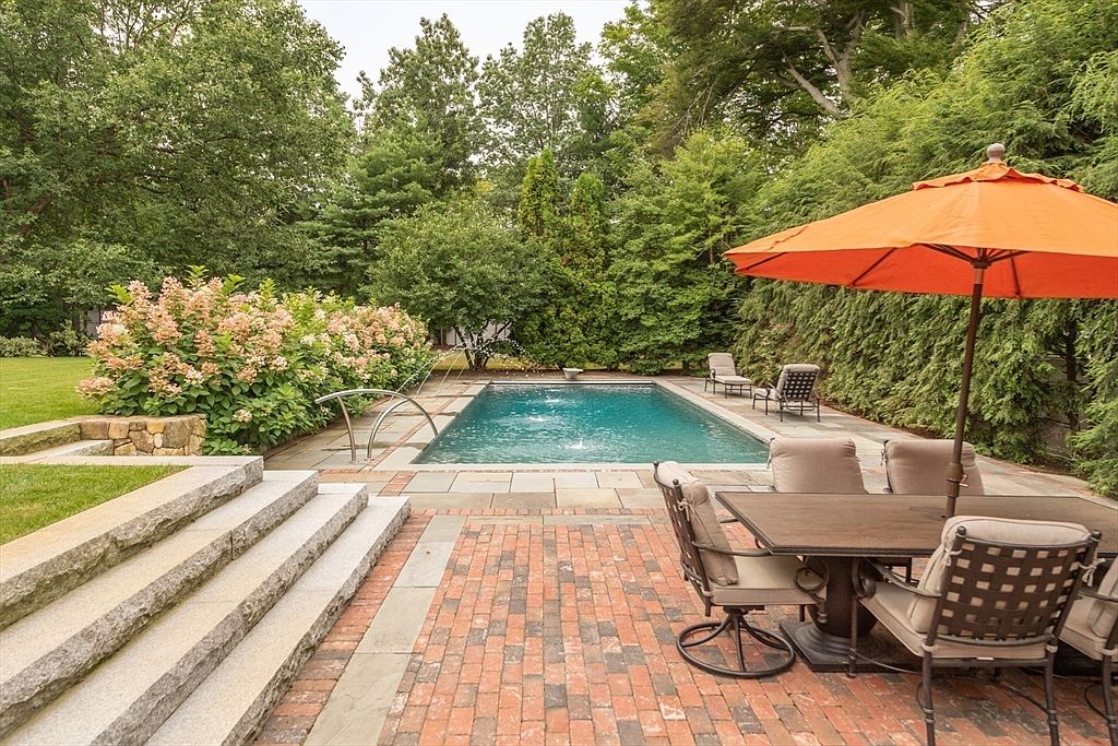 This inviting outdoor space features a rectangular swimming pool surrounded by a stone patio and a brick dining area. A large orange umbrella shades a dining set, while stone steps lead up to a lush lawn bordered by flowering shrubs and mature trees. The scene captures a serene, private backyard retreat perfect for summer entertaining.