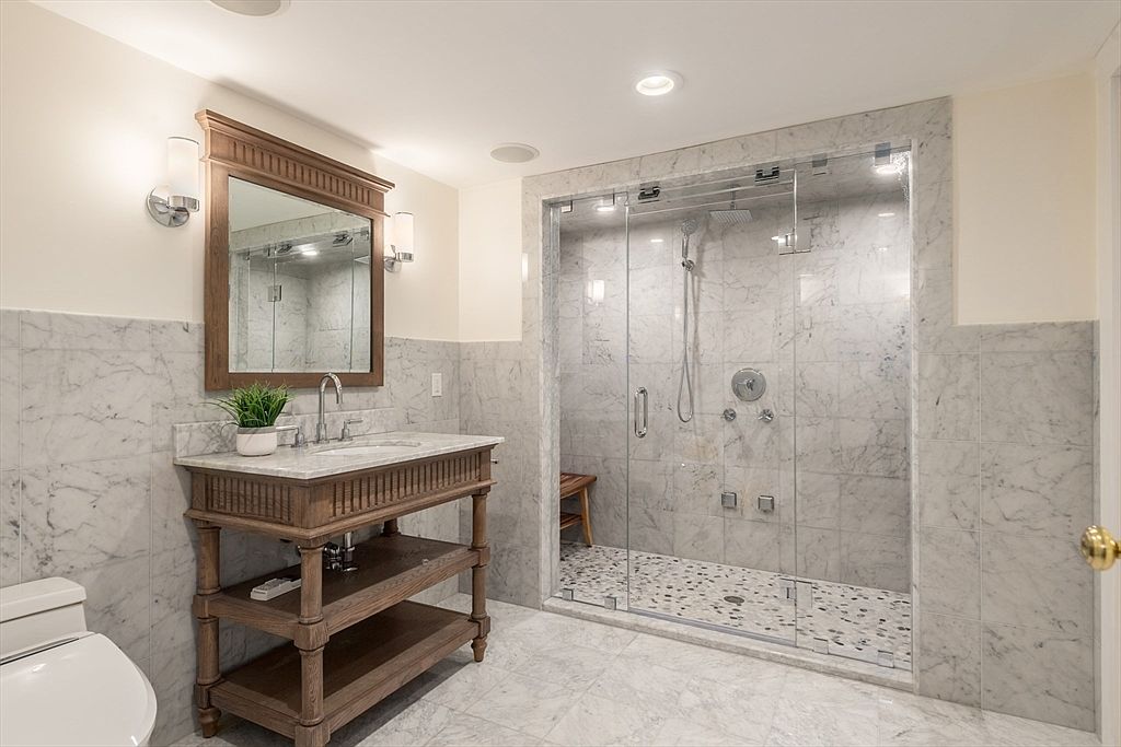 This elegant bathroom features a vanity with a stone countertop and a wooden base, paired with a matching framed mirror and wall-mounted light fixtures. To the right, a spacious walk-in shower is enclosed by glass panels and finished with stone wall tiles and a pebble-style floor. The room is accented with light-toned stone surfaces throughout, creating a cohesive and sophisticated aesthetic.