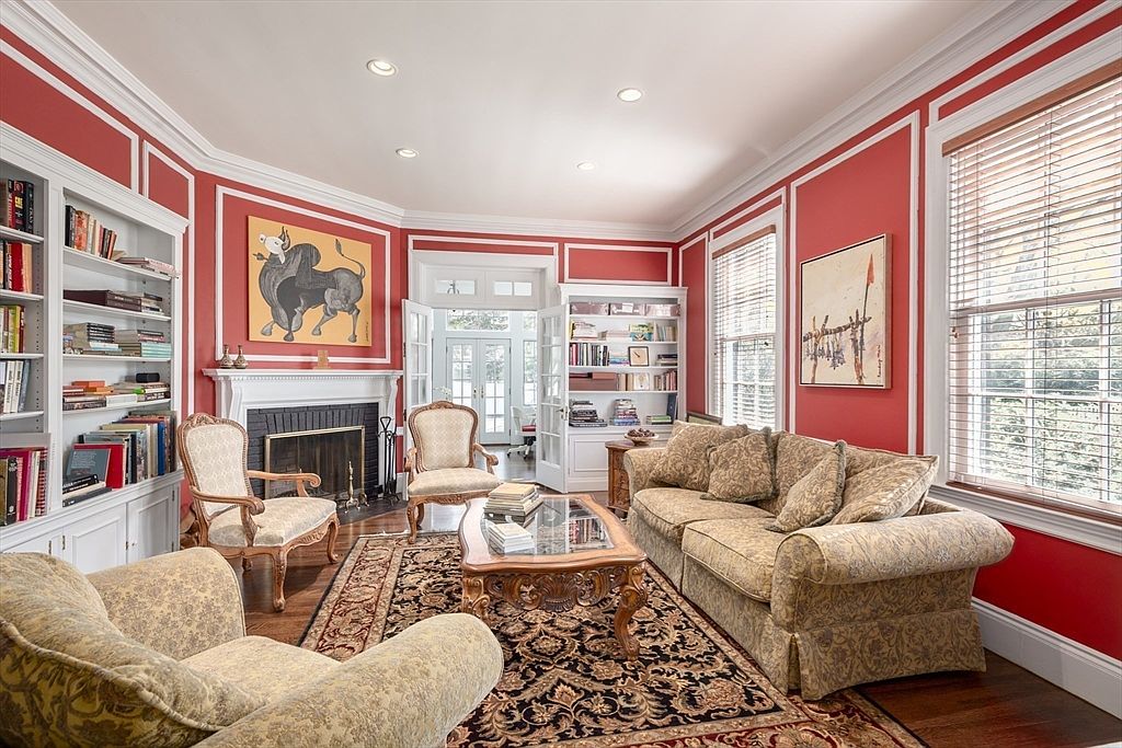This elegant living room features bold red walls accented by white wainscoting and crown molding, creating a sophisticated and traditional atmosphere. The space is anchored by a central fireplace with a classic mantel, flanked by built-in bookshelves, and furnished with a patterned sofa and matching armchairs arranged on a large, ornate area rug. Large windows with wooden blinds allow natural light to fill the room, which opens through double glass doors into an adjacent sunlit space.