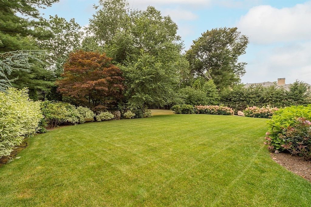 A lush, well-manicured lawn serves as the centerpiece of this expansive backyard, framed by mature trees and dense perimeter landscaping. Vibrant flowering shrubs add pops of color along the edges, while the soft, natural light suggests a peaceful, private setting. The perspective is a wide-angle view from the lawn looking toward the rear property line, emphasizing the generous outdoor space.