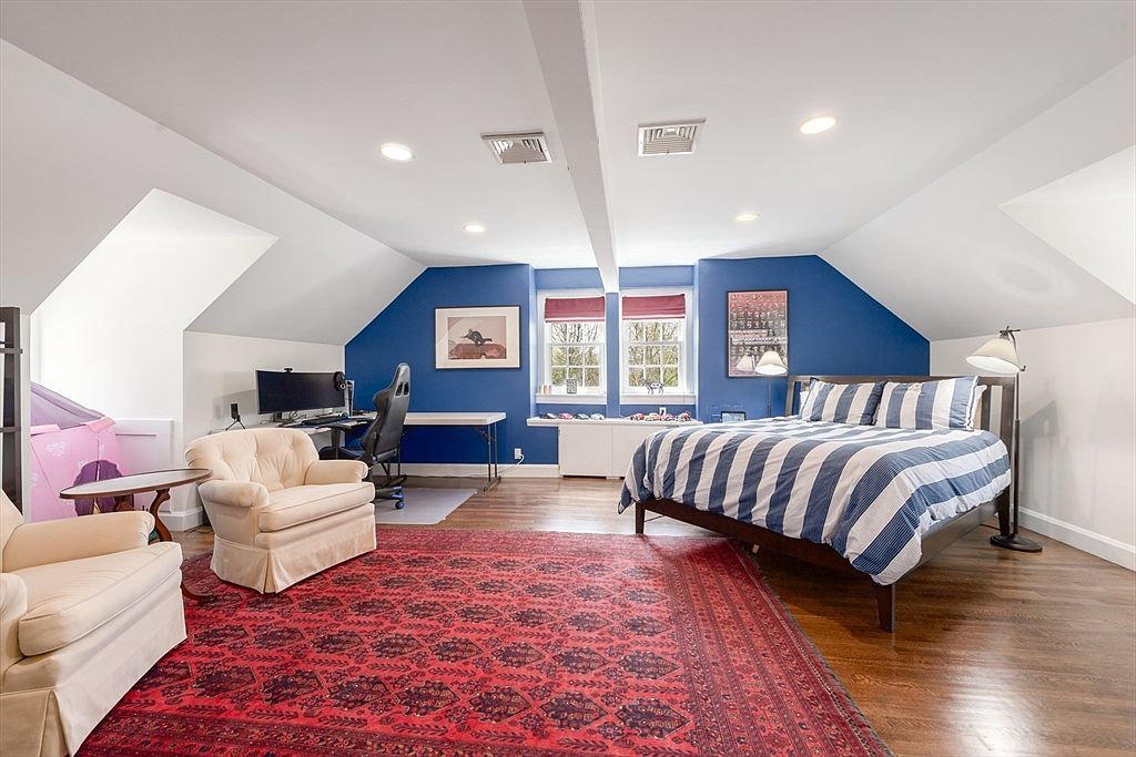 This spacious attic bedroom features a vaulted ceiling with recessed lighting and a striking blue accent wall. The room is furnished with a bed dressed in blue and white striped linens, a desk area, and two comfortable armchairs arranged on a large, ornate red patterned rug. The space is bright and airy, with dormer windows providing natural light and a warm, inviting atmosphere.