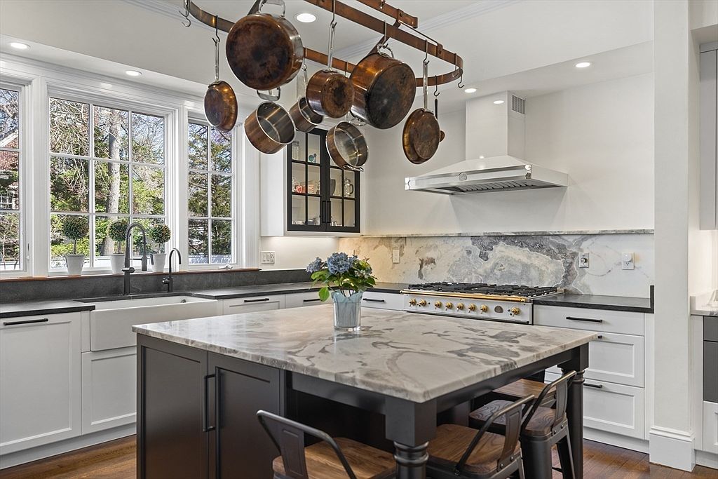 This elegant kitchen features a large central island with a stone countertop and dark cabinetry, complemented by white perimeter cabinets and a farmhouse sink. A prominent copper pot rack hangs above the island, while a professional-style range with a stainless steel hood serves as the focal point against a decorative stone backsplash. The space is brightened by large windows that provide a view of the outdoors, creating a sophisticated and functional culinary environment.