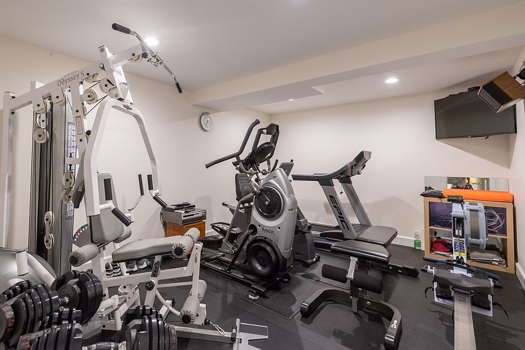 This home gym is equipped with a variety of fitness machines, including a multi-station weight system, a cross-trainer, a treadmill, and a rowing machine. The room features a dark rubberized floor, neutral-toned walls, and recessed lighting, creating a functional space for exercise. A wall-mounted television is positioned in the corner, and a small storage unit provides organization for workout accessories.