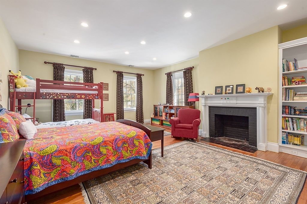 This spacious bedroom features a combination of a full-size bed with a vibrant, multi-colored patterned quilt and a bunk bed setup, making it ideal for a children's room. The room is anchored by a large, intricate area rug over hardwood floors and includes a traditional fireplace with a white mantel, a red armchair, and built-in bookshelves. Three windows with dark patterned curtains provide natural light, creating a warm and inviting atmosphere.