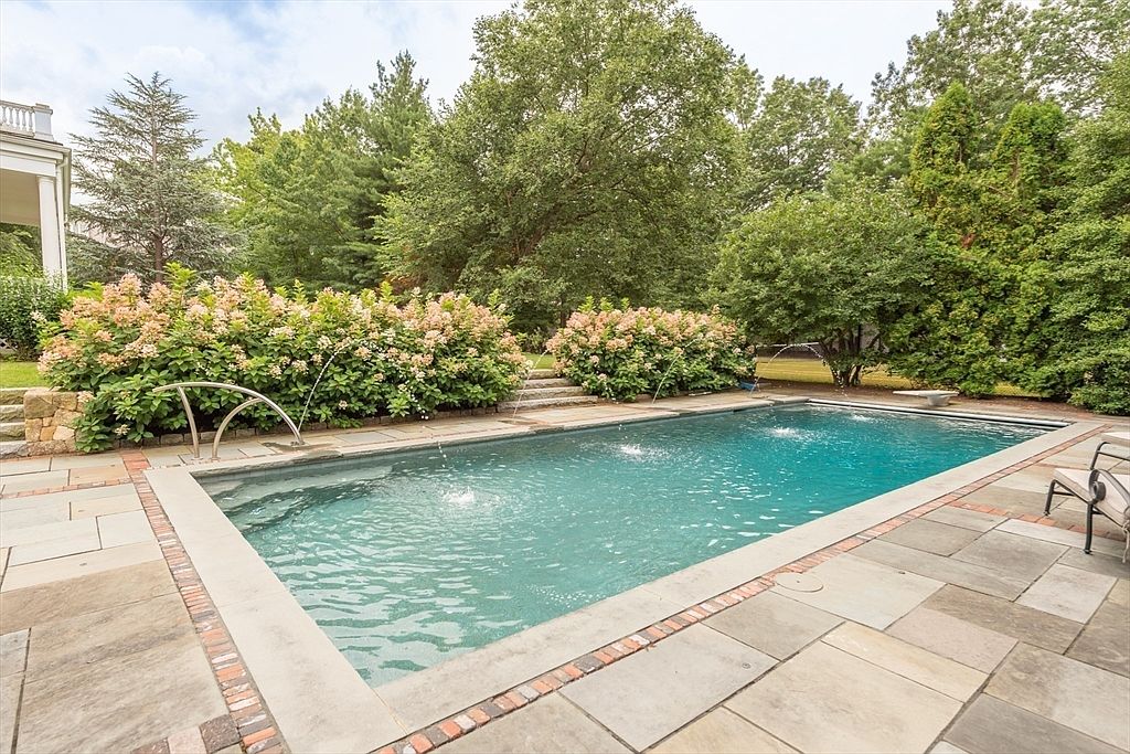 This backyard features a rectangular swimming pool with integrated water fountains, surrounded by a stone patio and lush landscaping. Large, blooming hydrangea bushes provide a soft, colorful border against the backdrop of mature trees. The scene is captured from a slightly elevated perspective, emphasizing the serene and private outdoor living space.