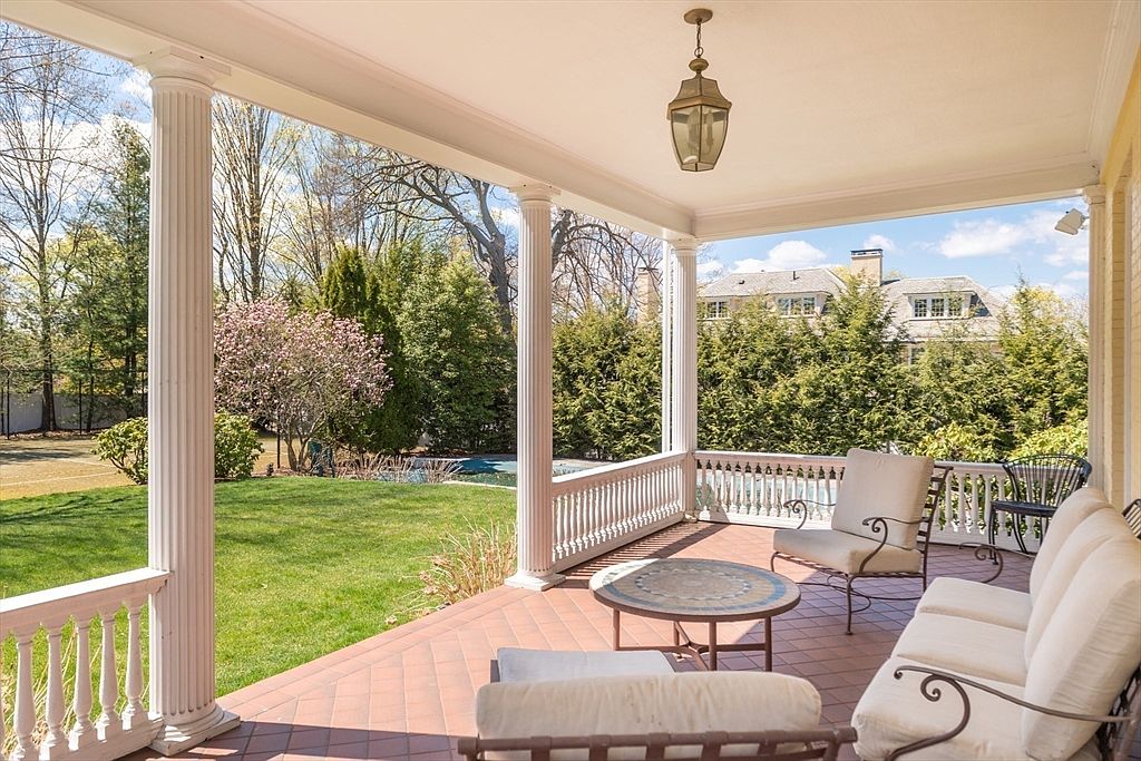This covered porch features a classic design with white columns and a matching railing, overlooking a lush green backyard. The space is furnished with comfortable outdoor seating, including a sofa and armchairs arranged around a circular tiled table. A hanging lantern provides overhead lighting, creating a serene and inviting atmosphere for outdoor relaxation.