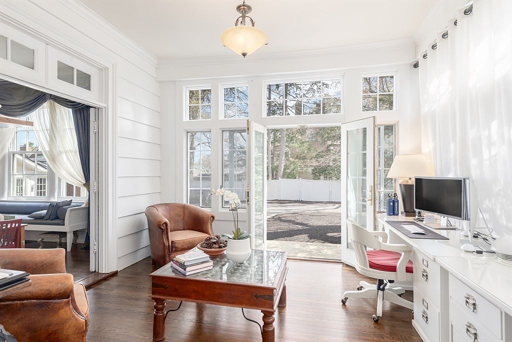 This bright and airy sunroom features white paneled walls, dark hardwood floors, and a central pendant light fixture. The space is furnished with two leather armchairs, a glass-topped coffee table, and a dedicated desk area with a computer setup. Large glass doors open to the outdoors, allowing ample natural light to fill the room and providing a seamless transition to the exterior.