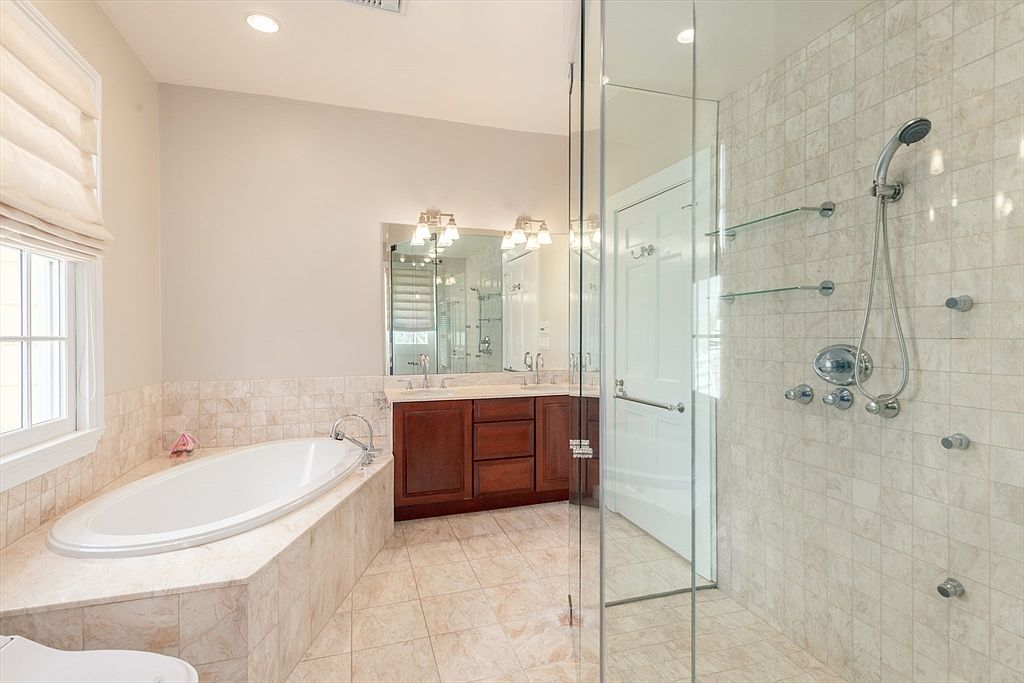This primary bathroom features a large soaking tub set into a tiled deck, positioned next to a window with soft fabric shades. A glass-enclosed shower stall with multiple wall-mounted fixtures and glass shelving sits to the right, while a double-sink vanity with dark wood cabinetry and a large mirror occupies the center wall. The space is finished with light-toned, patterned floor tiles and neutral wall paint, creating a bright and functional atmosphere.