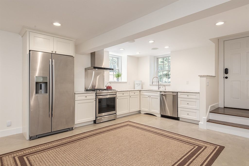 This bright, modern kitchen features white cabinetry, stainless steel appliances, and light-toned countertops. The space is open-concept with a neutral color palette, recessed lighting, and a large area rug defining the floor space. A set of stairs leads up to a door on the right, and two windows above the sink provide natural light to the workspace.
