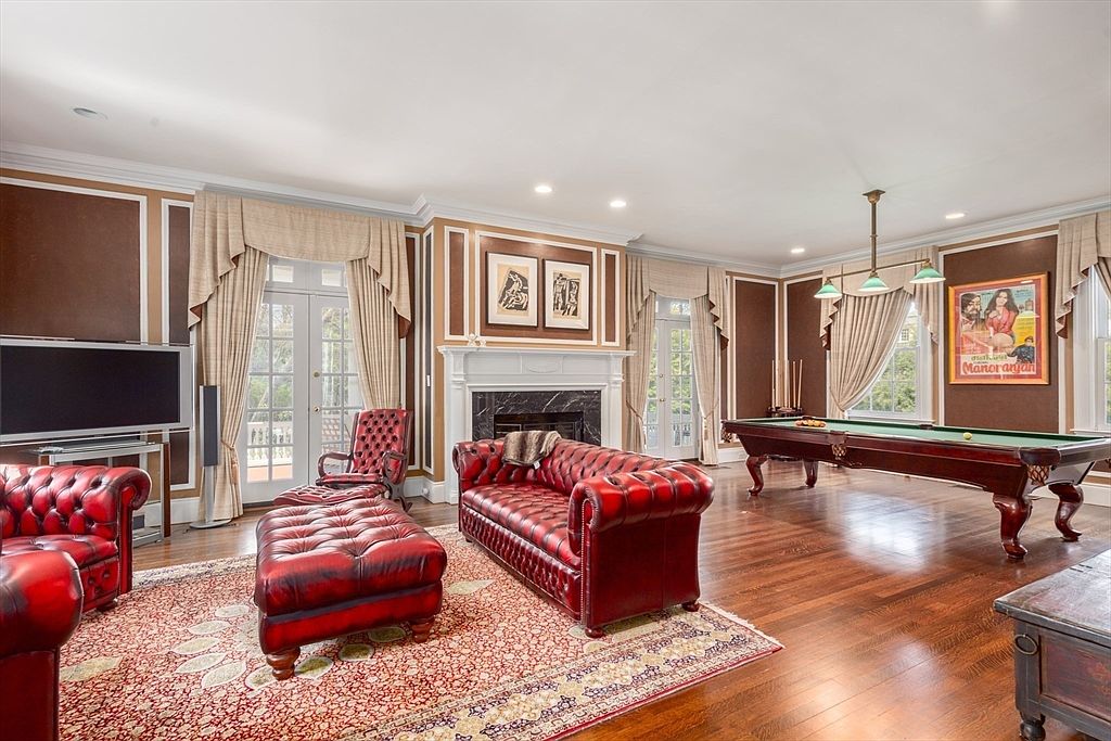 This spacious, traditional-style living room features rich dark wood paneling and hardwood flooring, anchored by a large, ornate fireplace with a mantel. The room is furnished with deep red leather seating, including a sofa and matching ottoman, and includes a full-sized billiard table positioned near large windows draped in elegant curtains. The perspective is a wide-angle shot from the corner of the room, capturing the blend of a formal lounge and a recreational space.