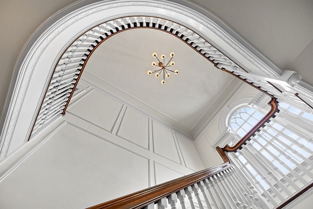 This low-angle, upward-looking perspective captures a grand, curved staircase featuring a polished wood handrail and classic white balusters. The space is defined by elegant architectural details, including intricate crown molding along the curved ceiling and paneled wall accents. A modern, multi-armed chandelier hangs centrally, casting a warm glow against the neutral, bright walls and highlighting the sophisticated, traditional design of the foyer.