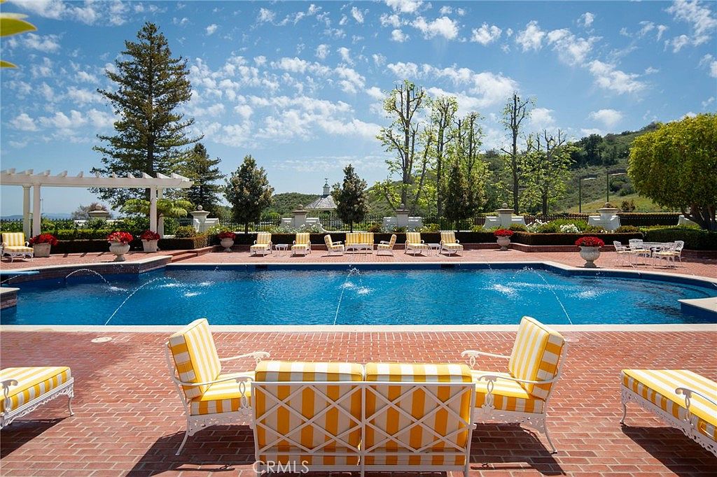 This expansive outdoor space features a large, rectangular swimming pool as the centerpiece, surrounded by a classic red brick patio. The area is furnished with elegant white metal patio furniture featuring yellow and white striped cushions, creating a bright and inviting atmosphere. In the background, lush green trees, manicured hedges, and a distant gazebo set against a blue sky with scattered clouds provide a serene and luxurious estate-like setting.