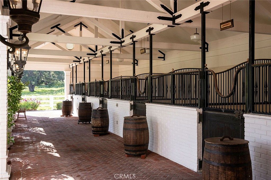 This image showcases a sophisticated, covered equestrian stable aisle featuring a classic brick floor and white-painted brick stall bases. The space is lined with dark metal stall doors and accented by rustic wooden barrels placed in front of each stall, creating a refined, country-estate aesthetic. The perspective looks down the length of the open-air corridor, highlighting the clean lines of the timber-framed ceiling and the peaceful, sunlit outdoor view at the far end.