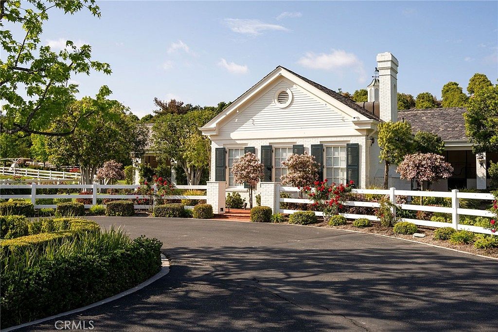 This charming, white-painted brick home features a classic gabled roofline and symmetrical windows with dark shutters, exuding a timeless, elegant aesthetic. A curved asphalt driveway leads up to the property, framed by a pristine white picket fence, manicured hedges, and vibrant rose bushes. The scene is captured from a low-angle perspective, emphasizing the welcoming curb appeal and the lush, well-maintained landscaping.