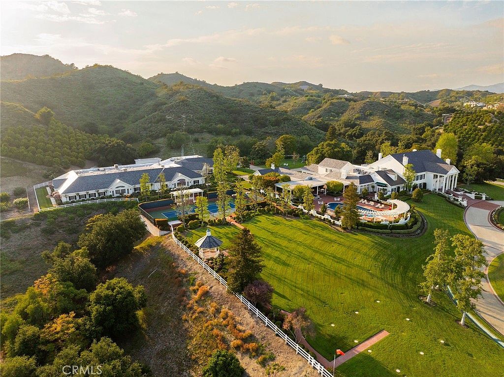 This expansive aerial view captures a luxurious estate nestled within rolling, sun-drenched hills. The property features a sprawling main residence with a classic architectural style, a large manicured lawn, a private tennis court, and a resort-style swimming pool area. The cinematic perspective highlights the vastness of the grounds and the serene, secluded nature of this high-end residential setting.