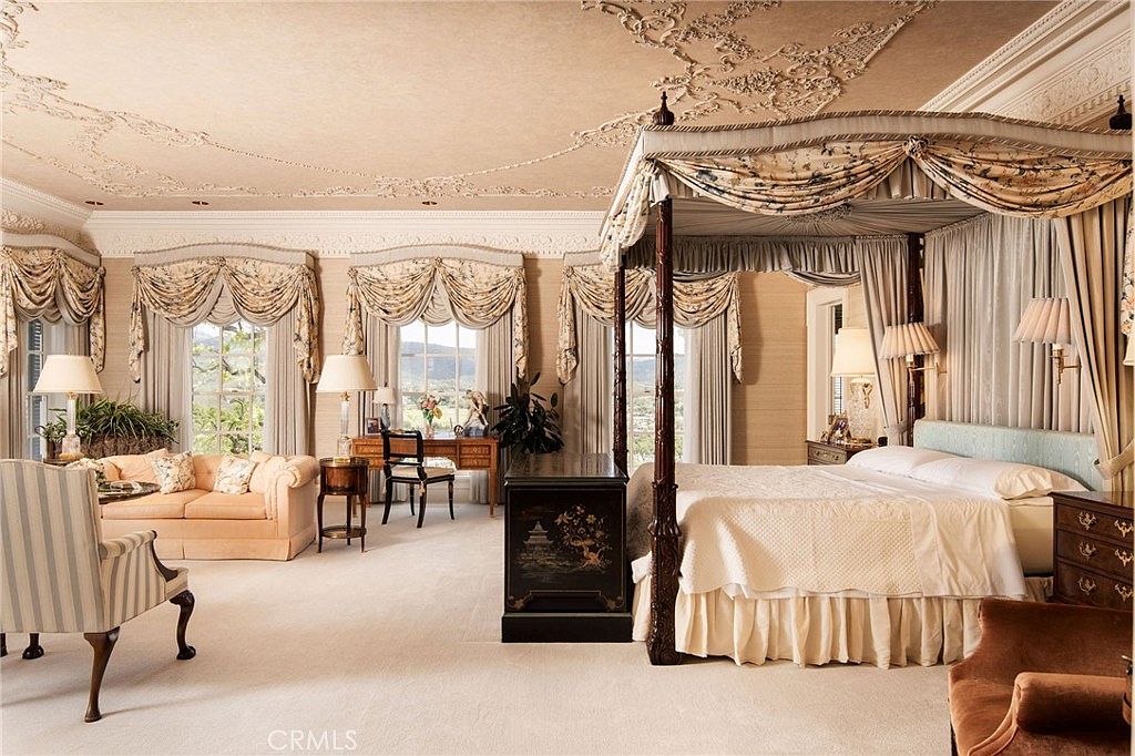 This opulent primary bedroom features a grand four-poster canopy bed as its centerpiece, complemented by intricate ceiling molding and elegant drapery. The room includes a cozy seating area with a plush sofa, a dedicated desk space, and large windows that invite natural light while offering scenic views. The overall aesthetic is one of classic luxury and sophisticated comfort, perfect for a high-end estate.