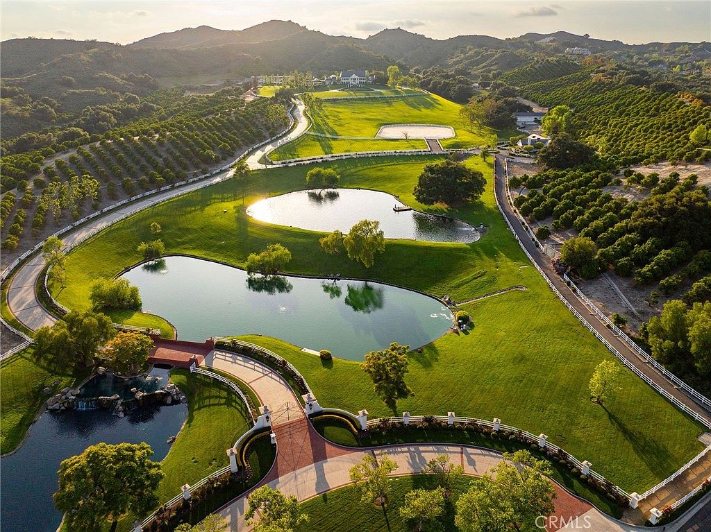 This stunning aerial view captures a sprawling, manicured estate featuring multiple serene ponds, lush green lawns, and extensive orchards. A winding driveway leads through the property, which is framed by rolling hills and mature trees, creating an atmosphere of luxury and tranquility. The perspective highlights the vast scale and meticulous landscaping of this private countryside retreat.