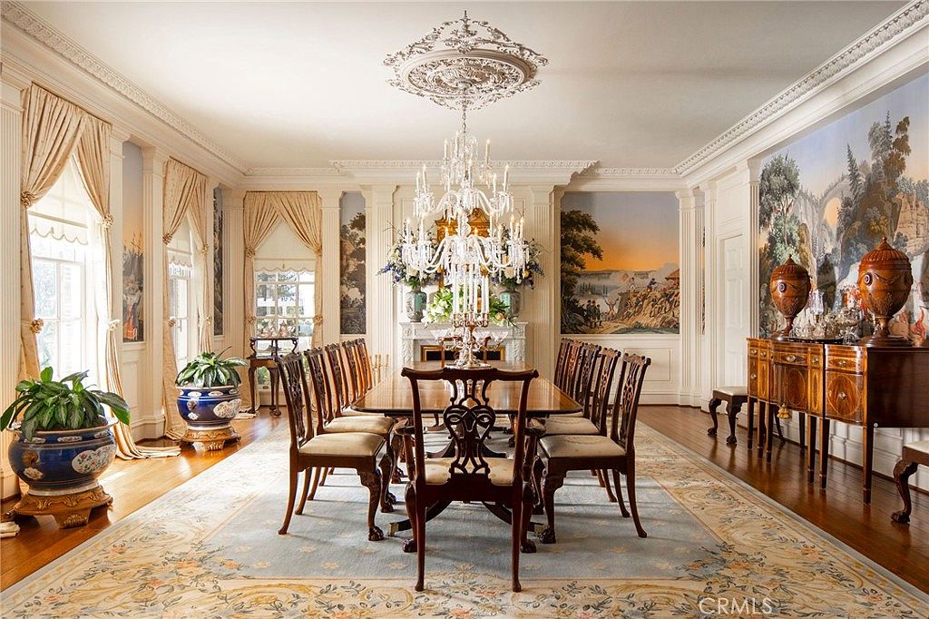 This formal dining room exudes elegance with its classic architectural details, including intricate crown molding, a grand crystal chandelier, and scenic wall murals. A long, polished wooden table is surrounded by matching chairs, all set atop a large, ornate area rug that anchors the space. The room is bathed in natural light from tall windows draped in soft curtains, creating a sophisticated and luxurious atmosphere perfect for hosting formal gatherings.