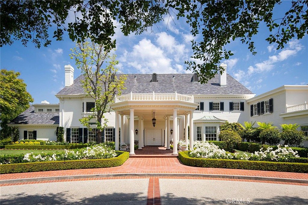 This grand, traditional-style estate features a symmetrical white brick facade with a prominent, columned portico serving as the main entryway. The property is framed by lush, manicured landscaping, including vibrant green hedges and blooming white rose bushes, all set against a clear blue sky. A brick-paved driveway leads directly to the stately entrance, creating an inviting and prestigious first impression.