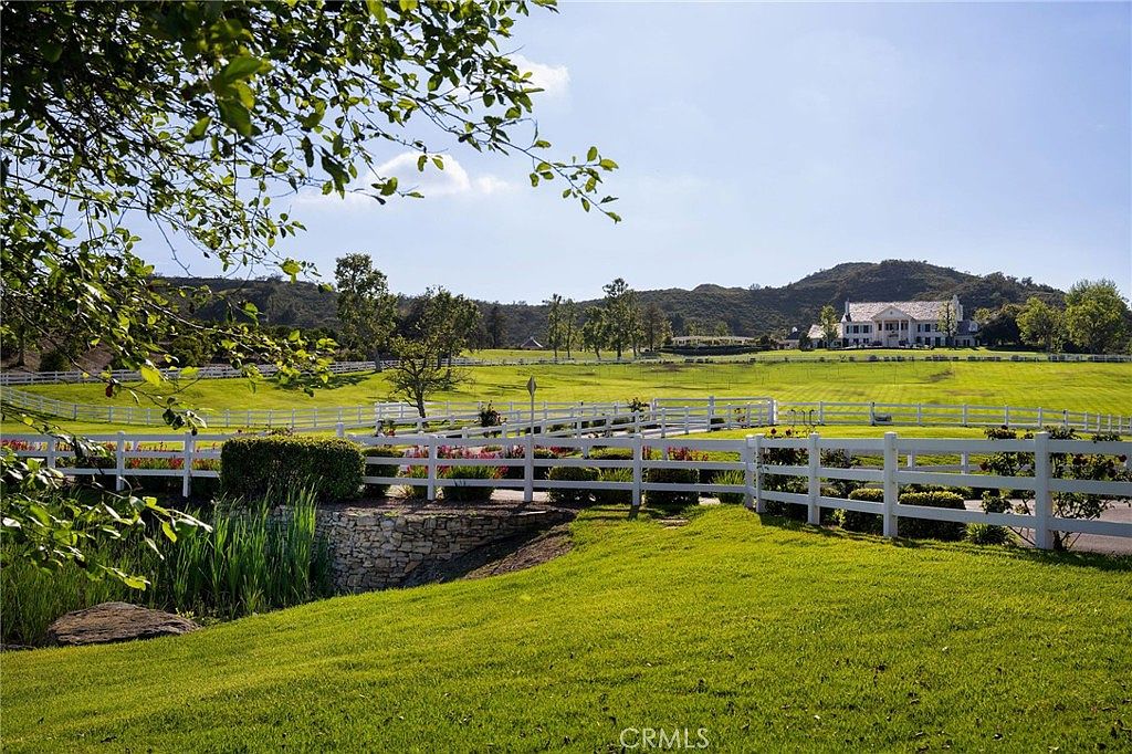 This expansive estate features a sprawling, manicured green lawn enclosed by a classic white rail fence, leading the eye toward a grand, traditional-style mansion in the distance. The foreground includes a stone-walled garden bed with vibrant flowers and a lush grassy slope, creating a serene and picturesque pastoral setting. The perspective is a wide, eye-level shot that emphasizes the vastness of the property and its tranquil, park-like atmosphere.