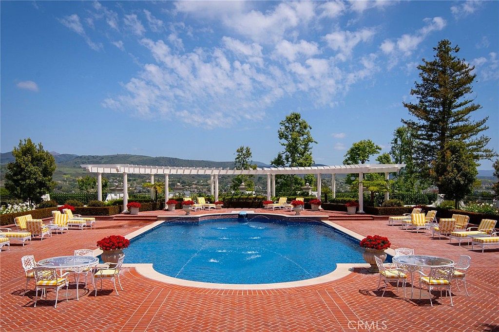 This expansive outdoor pool area features a large, custom-shaped swimming pool with integrated fountains, surrounded by a classic red brick patio. A long, white pergola structure provides architectural interest and shade, while numerous sets of patio furniture offer ample space for lounging and dining. The setting is framed by lush greenery and distant mountain views, creating a luxurious and serene atmosphere.