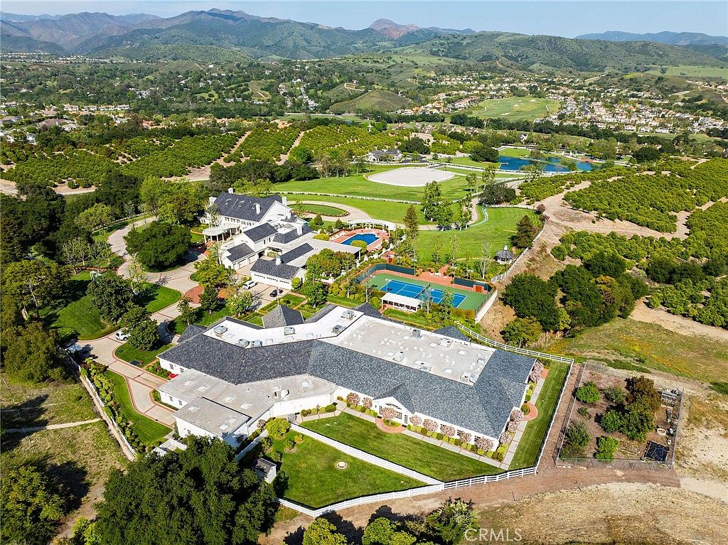 This expansive aerial view captures a sprawling luxury estate set against a backdrop of rolling hills and mountains. The property features a massive main residence with a complex roofline, a private tennis court, a swimming pool, and extensive manicured grounds surrounded by lush orchards. The perspective emphasizes the grand scale and secluded, resort-like nature of this impressive countryside compound.
