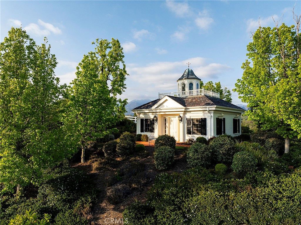 This elegant, white-painted structure features a classic architectural design with a prominent central cupola and a symmetrical facade. Surrounded by lush, manicured greenery and mature trees, the building sits atop a gentle rise, offering a picturesque and serene aesthetic. The perspective is captured from a slightly elevated angle, emphasizing the building's stately presence against a clear blue sky.
