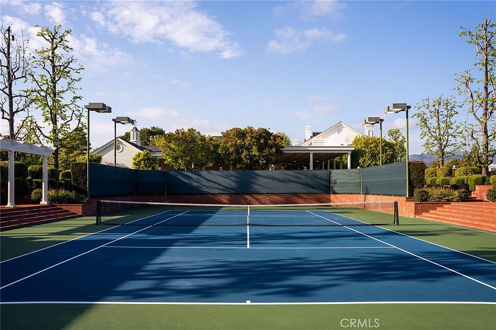 This image features a well-maintained, professional-grade tennis court with a vibrant blue playing surface and a crisp white net. The court is enclosed by a dark green privacy fence and brick retaining walls, with tall stadium-style lighting fixtures positioned around the perimeter. In the background, lush green trees and a glimpse of a classic white building suggest a serene, upscale residential or community setting.