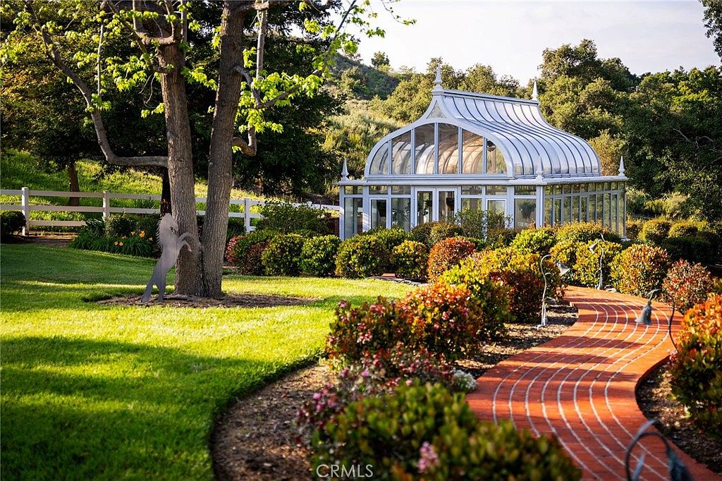 This picturesque garden scene features a stunning, Victorian-style glass conservatory nestled among lush greenery and rolling hills. A curved brick pathway leads the eye through manicured landscaping and vibrant bushes toward the structure, creating a serene and elegant atmosphere. The perspective is a ground-level shot that emphasizes the peaceful, park-like setting of the property.