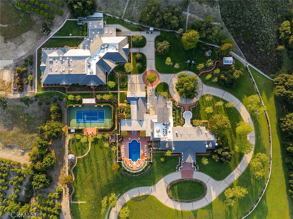 This stunning aerial view captures a sprawling luxury estate set within a lush, manicured landscape. The property features a large main residence with multiple wings, a private tennis court, and a classic rectangular swimming pool surrounded by expansive green lawns and mature trees. The cinematic perspective highlights the grand scale of the property, including its intricate driveway layout and surrounding agricultural or orchard land.