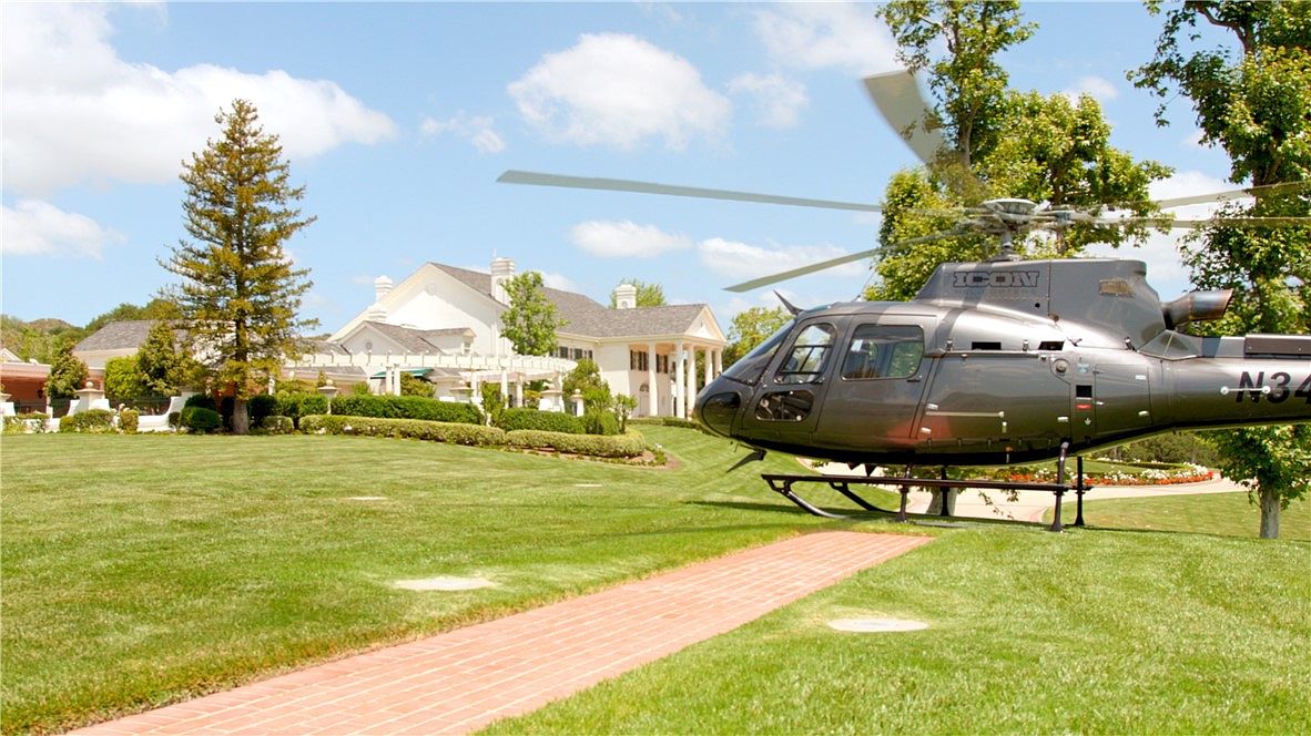 This expansive estate features a meticulously manicured lawn and a brick pathway leading toward a grand, white-columned residence. A dark-colored helicopter is prominently parked on the grass in the foreground, adding a sense of luxury and exclusivity to the property. The scene is captured from a low-angle perspective, emphasizing the scale of the grounds and the impressive architectural details of the home.