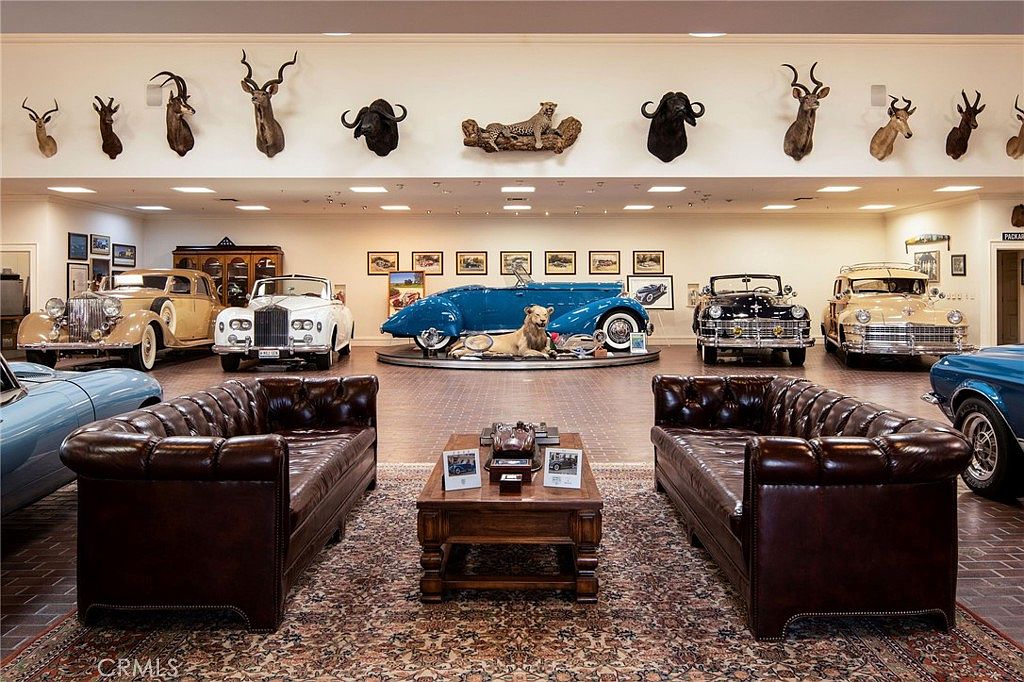 This expansive, museum-like garage space showcases a collection of vintage luxury automobiles arranged around a central seating area featuring two large, tufted leather sofas and a wooden coffee table. The walls are adorned with numerous taxidermy animal mounts and framed automotive photographs, creating a sophisticated and eclectic atmosphere. The perspective is a wide, symmetrical shot from the center of the room, emphasizing the grand scale and curated nature of this private car gallery.