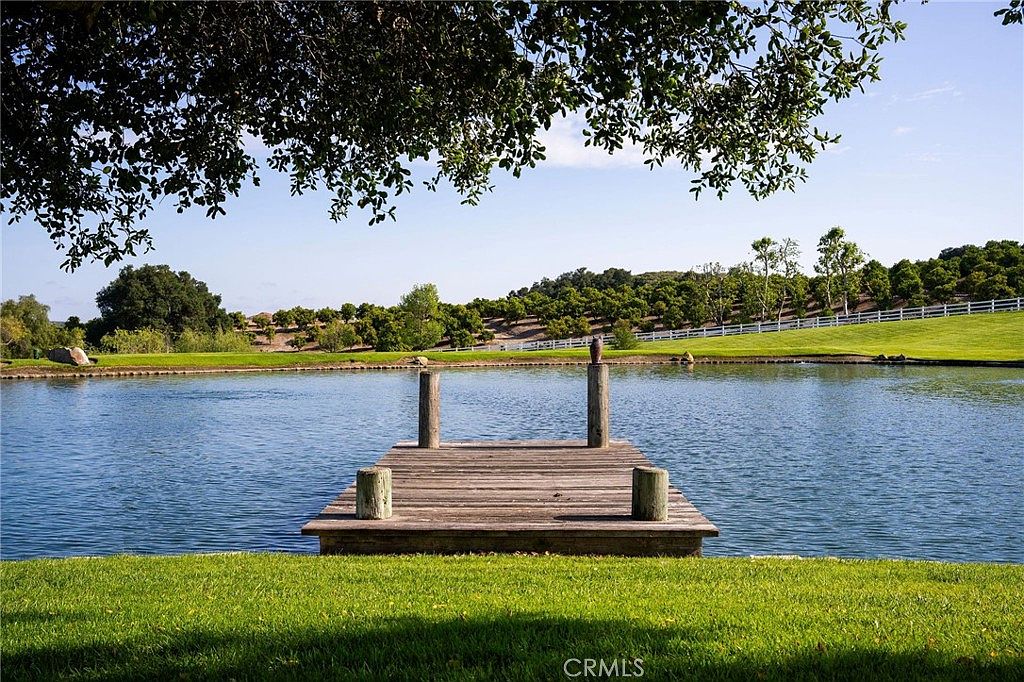 This serene outdoor scene features a wooden dock extending into a calm, reflective pond, framed by lush green grass and a large, overhanging tree in the foreground. In the background, a gentle hillside is covered in rows of trees, creating a peaceful and picturesque landscape. The perspective is eye-level, capturing the tranquil beauty of the property's natural water feature.