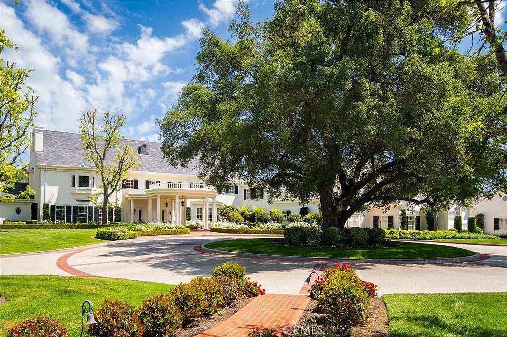 This grand estate features a classic, stately architectural style with a prominent circular driveway and a large, mature oak tree serving as a focal point in the front yard. The white exterior is accented by dark shutters and a multi-gabled roof, leading to an elegant covered entryway with columns. The meticulously manicured landscaping and brick-lined pathways create a sophisticated and welcoming first impression.