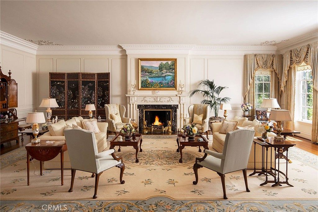 This elegant and formal living room features a symmetrical layout centered around a grand fireplace with a marble surround and a landscape painting above. The space is furnished with plush, light-toned seating, ornate side tables, and multiple lamps, all set upon a large, intricate area rug. The room exudes a sophisticated, traditional aesthetic with detailed wall paneling, crown molding, and floor-to-ceiling drapery framing the windows.