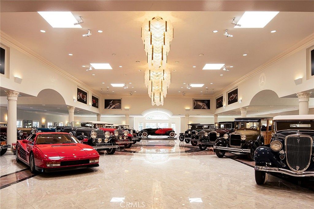 This expansive, high-end private garage functions as a luxurious showroom for a collection of classic and exotic automobiles. The space features polished marble flooring, grand crystal chandeliers, and recessed lighting, creating an opulent atmosphere that highlights the vehicles. The wide-angle perspective captures the symmetry of the room, emphasizing the scale and prestige of this unique automotive display area.