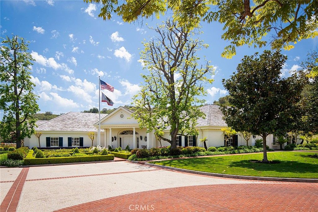 This grand, single-story estate features a classic architectural style with a light-colored brick facade and a prominent, columned entryway. The property is framed by lush, manicured landscaping, mature trees, and a wide, circular brick-accented driveway that leads to the main entrance. The scene is captured from a low-angle, eye-level perspective, emphasizing the home's stately presence and expansive, well-maintained grounds under a bright, clear sky.
