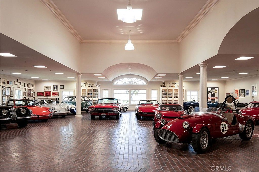 This expansive, high-end indoor garage functions as a private showroom for a collection of vintage and classic sports cars. The space features a polished brick floor, high ceilings with recessed lighting, and white columns, creating an elegant and museum-like atmosphere. The wide-angle perspective captures the impressive scale of the room, highlighting the pristine condition of the vehicles on display.