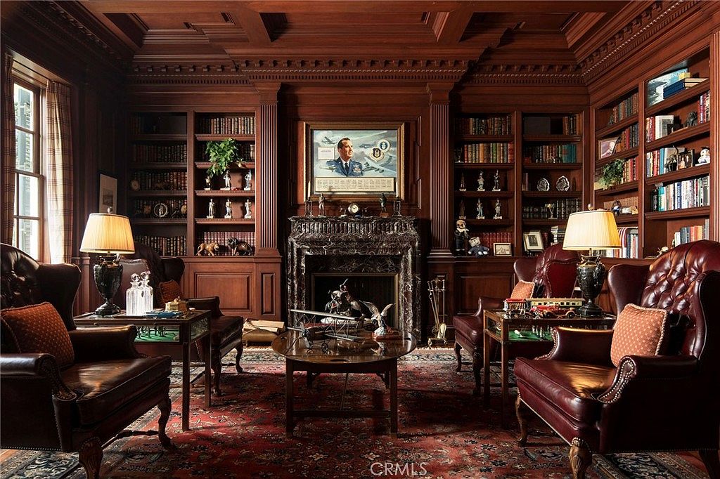 This sophisticated, traditional-style study features rich, dark wood paneling, built-in floor-to-ceiling bookshelves, and a grand marble fireplace as the central focal point. The room is furnished with plush, tufted leather armchairs, ornate side tables, and a central coffee table displaying aviation-themed decor, all set atop a classic, intricate Persian-style rug. The warm, ambient lighting creates an atmosphere of scholarly elegance and timeless luxury.