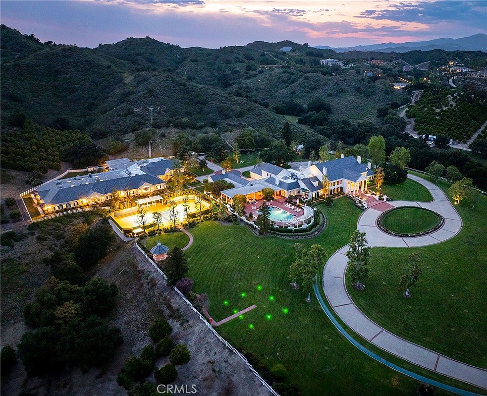 This stunning aerial view captures a sprawling luxury estate nestled against a backdrop of rolling hills at dusk. The property features a large main residence, a separate structure with a lighted tennis court, a resort-style swimming pool, and expansive manicured lawns connected by a winding driveway. The warm glow of the interior and landscape lighting creates an inviting and sophisticated atmosphere, highlighting the estate's grand scale and private setting.