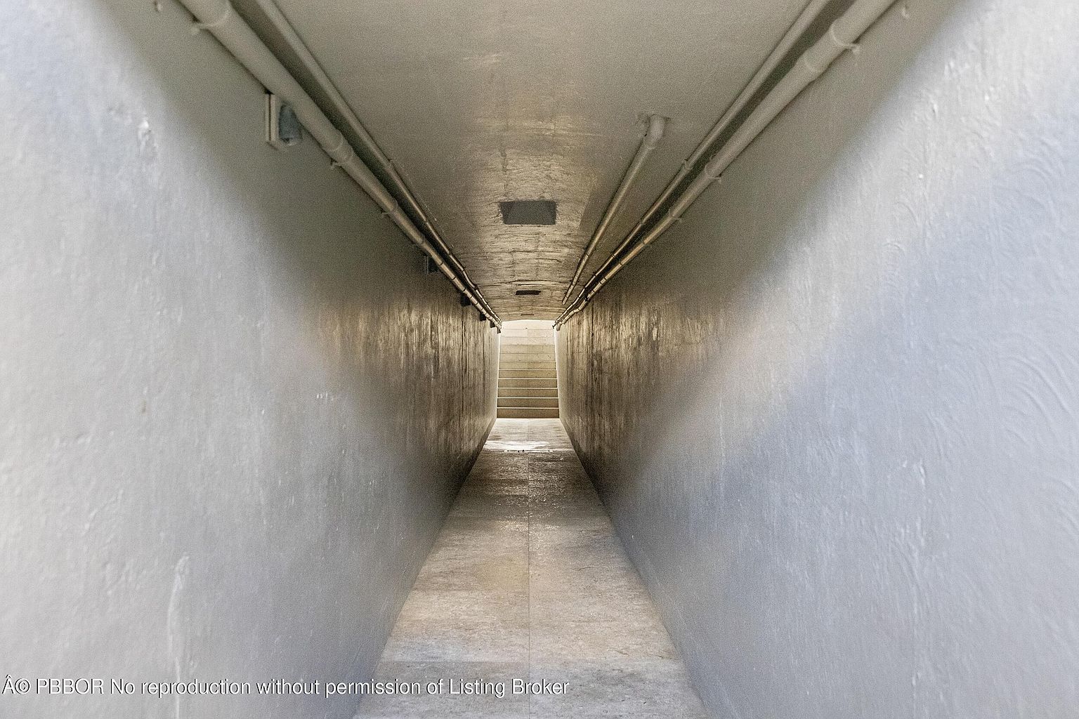 This image captures a long, narrow, enclosed corridor leading to a set of stairs at the far end. The space features smooth, light-colored walls and a tiled floor, with exposed pipes running along the ceiling. The perspective is centered and symmetrical, creating a tunnel-like effect that draws the eye toward the illuminated staircase.