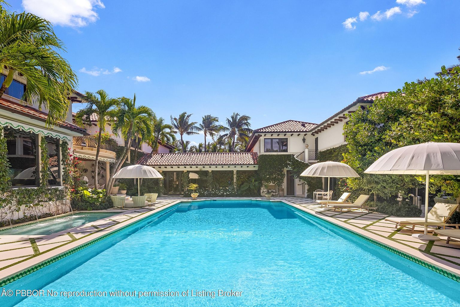 This expansive Mediterranean-style estate features a large, rectangular swimming pool as the centerpiece of a private courtyard. The surrounding patio is paved with geometric stone tiles, complemented by lush greenery, palm trees, and multiple seating areas shaded by large umbrellas. The architecture showcases white stucco walls, terracotta roof tiles, and climbing vines, creating a serene and luxurious outdoor living environment.